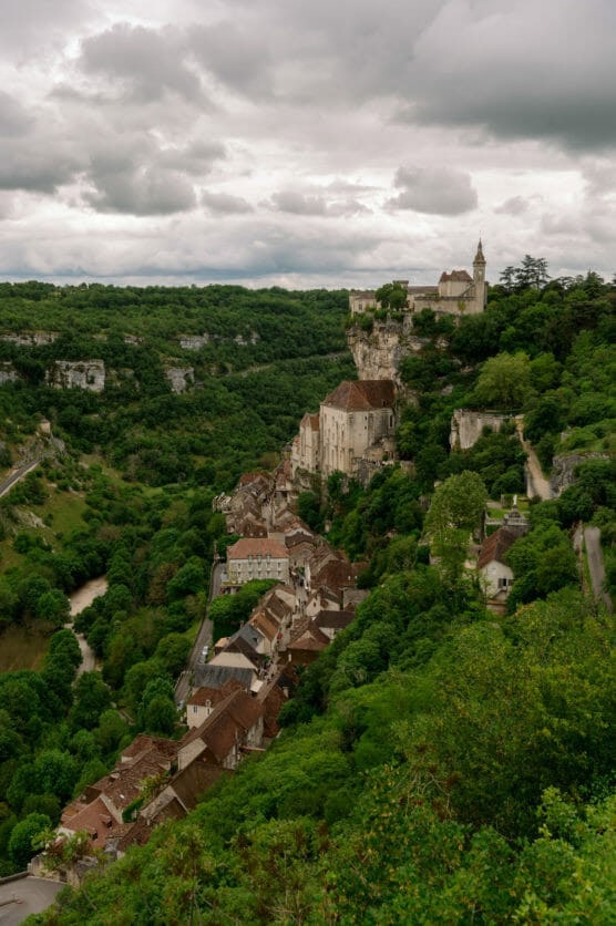 A hillside village with red-roofed houses surrounded by lush green trees, with a church and buildings perched on a rocky cliff under a cloudy sky. by Justin Salem Meyer