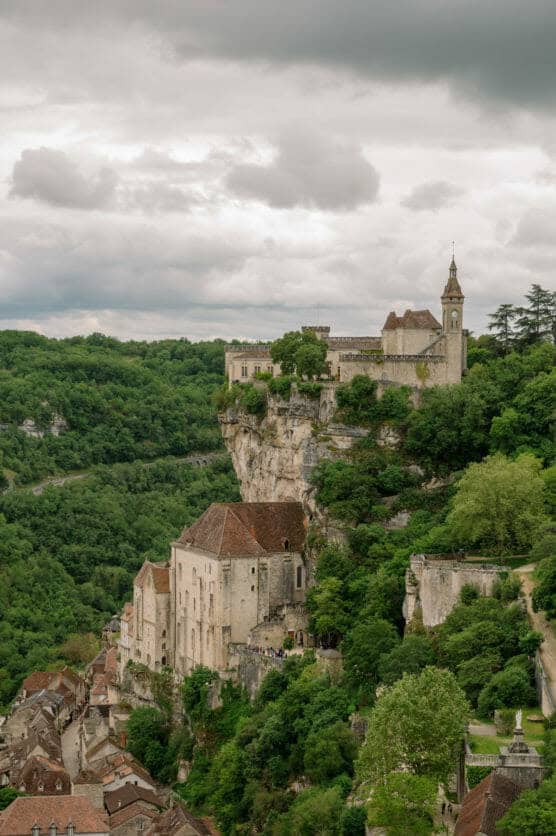 A historic village with stone buildings and a church sits atop a steep, green cliffside, surrounded by lush trees under a cloudy sky. by Justin Salem Meyer