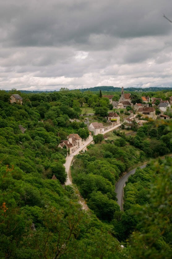 A winding road leads through a lush green valley to a small village with stone houses and a church, all surrounded by dense trees under a cloudy sky. by Justin Salem Meyer