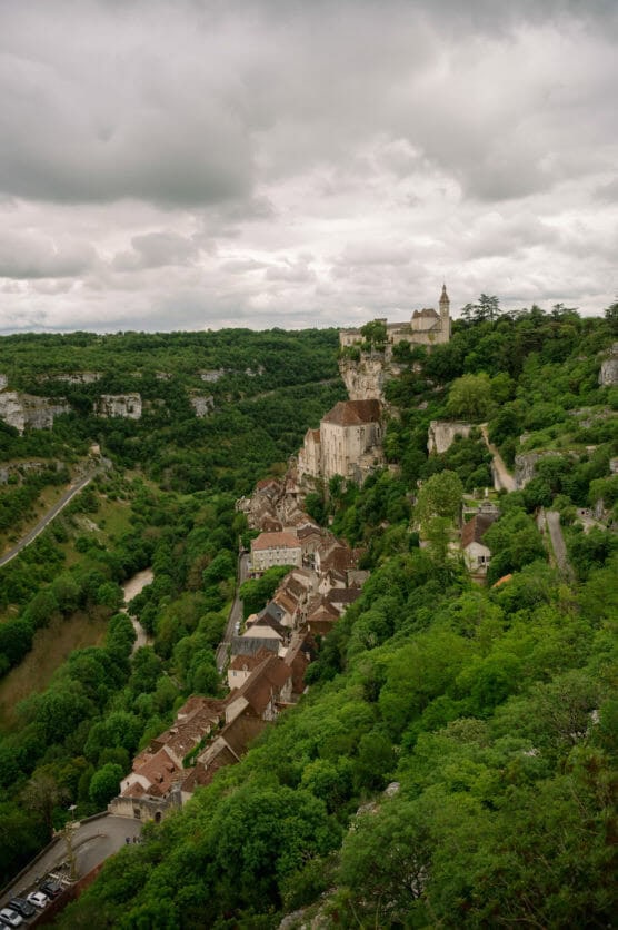 A hilltop village with stone buildings and a church is nestled among lush green trees and cliffs under a cloudy sky, overlooking a winding road and river below. by Justin Salem Meyer