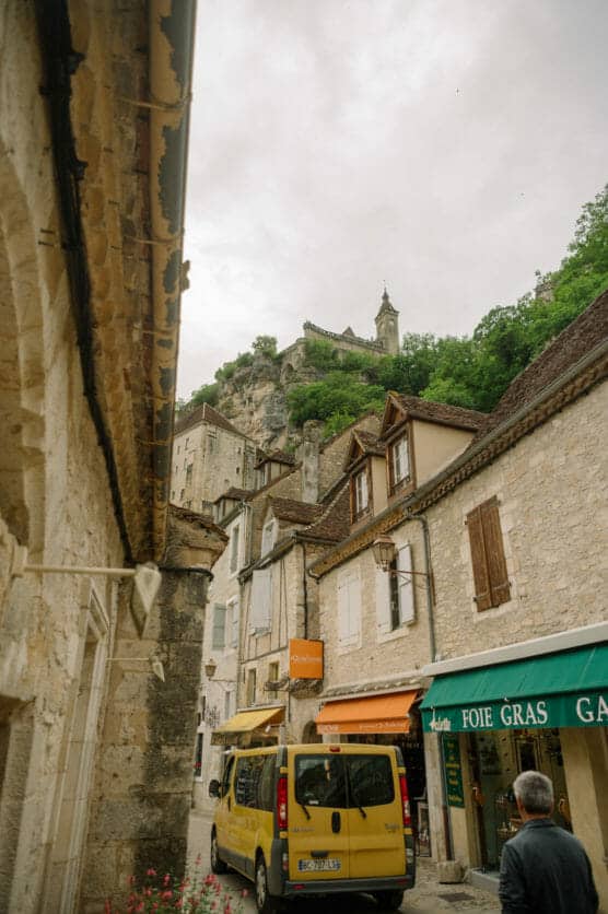 A narrow stone street in a historic village, with a yellow van parked outside shops and a person walking. Above, a church sits atop a rocky hill surrounded by greenery under a cloudy sky. by Justin Salem Meyer