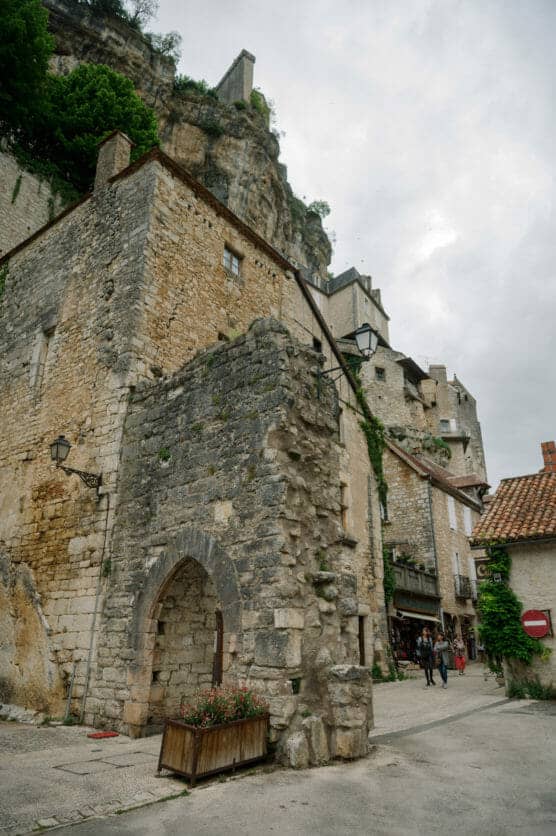 Stone buildings with arched doorways and ivy line a narrow street in a historic village at the base of a rocky cliff. A few people walk along the street under a cloudy sky. by Justin Salem Meyer