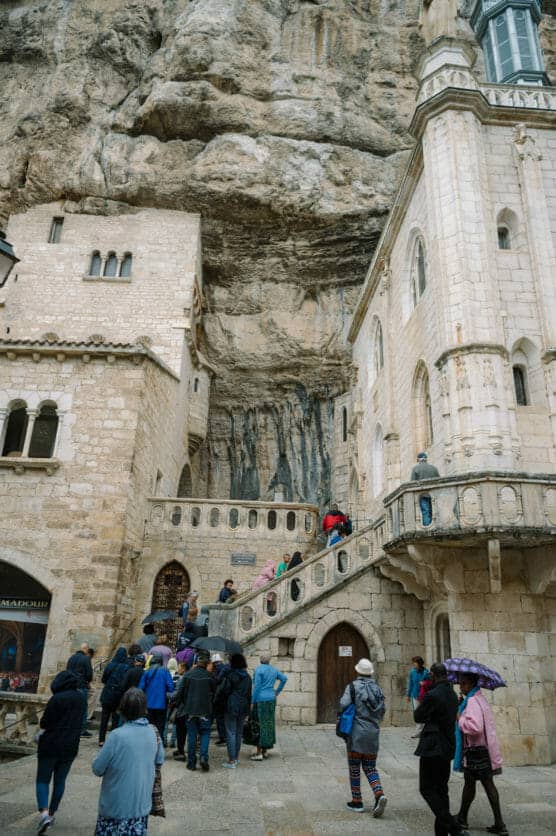 A group of people with umbrellas stands and walks near stone buildings built into a rocky cliff, with some people ascending an outdoor staircase. The architecture features arched windows and decorative stonework. by Justin Salem Meyer