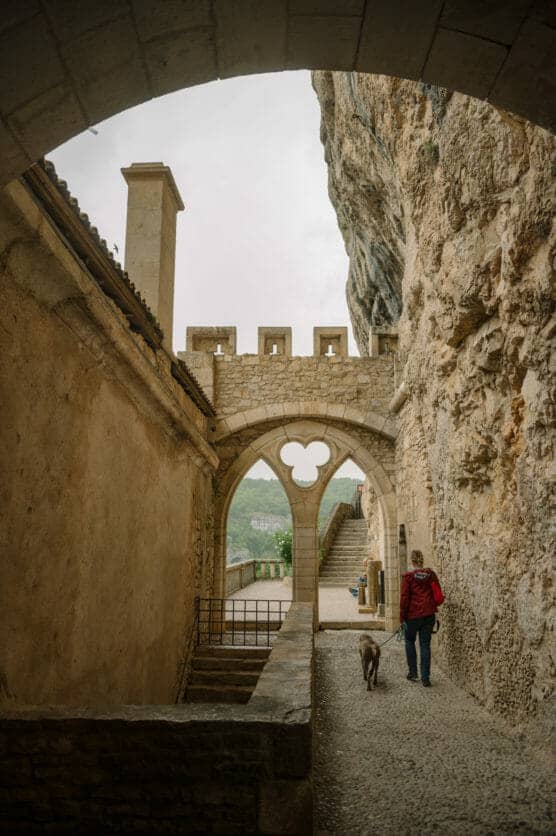 A person in a red jacket walks a dog along a stone pathway next to a historic building and rock wall, passing under a Gothic-style stone archway with a view of stairs and greenery beyond. by Justin Salem Meyer