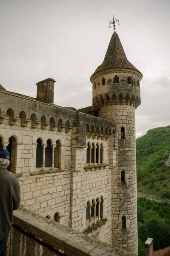 A stone castle with arched windows and a round tower topped with a pointed roof stands on a hillside. A person with a hat is seen in the foreground near a railing. Lush green trees fill the background. by Justin Salem Meyer