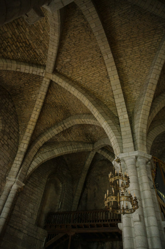 A vaulted stone ceiling with intersecting arches in a historic building, possibly a church; a brass chandelier hangs in the foreground and a wooden balcony is visible along the back wall. by Justin Salem Meyer