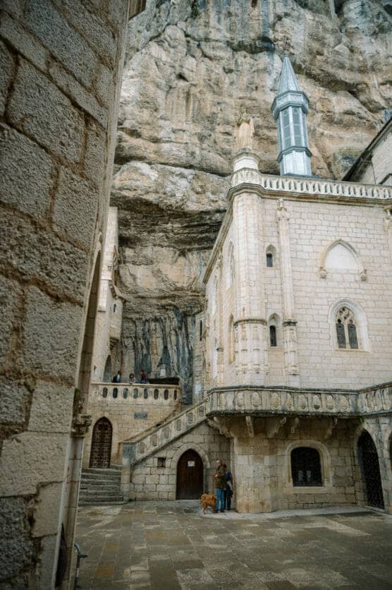A stone building with arched windows and a tower is built into a rocky cliff. A person and a dog stand in the courtyard below, and a few people are visible on the steps and balcony above. by Justin Salem Meyer