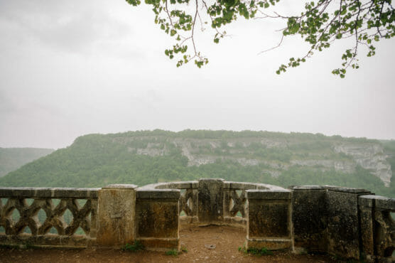A stone viewpoint with a weathered railing overlooks a lush, green valley and distant rocky cliffs under an overcast sky. Tree branches with green leaves frame the top of the image. by Justin Salem Meyer