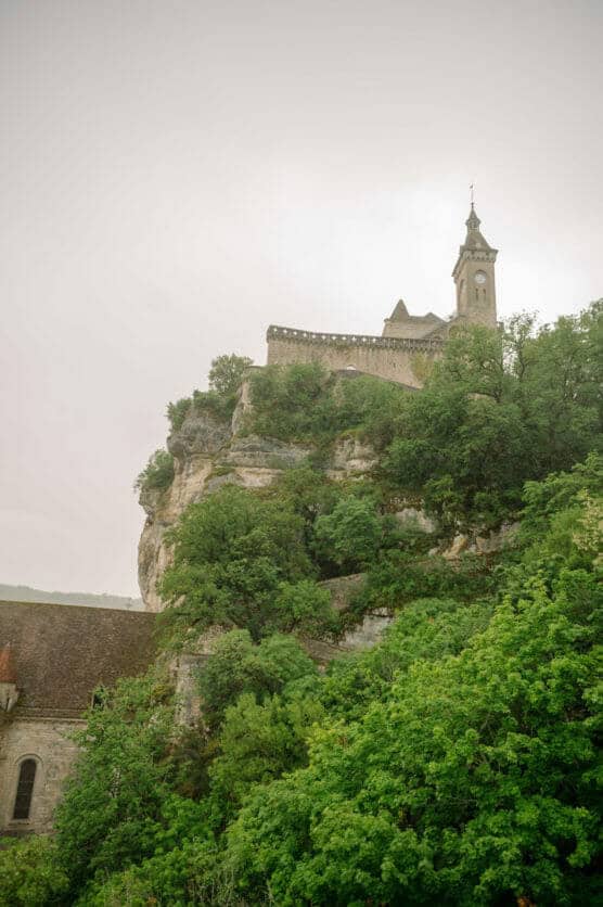 A stone church with a tall steeple sits atop a rocky cliff surrounded by lush green trees, under a cloudy sky. by Justin Salem Meyer