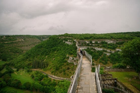 A person stands on a stone walkway with metal railings, overlooking lush green hills and a winding path below, under a cloudy sky. by Justin Salem Meyer