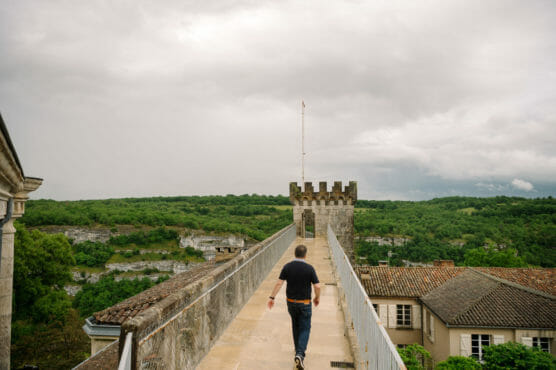 A person walks along a stone walkway toward a small castle tower, with green hills and cloudy skies in the background. Rooftops of nearby buildings are visible to the right. by Justin Salem Meyer