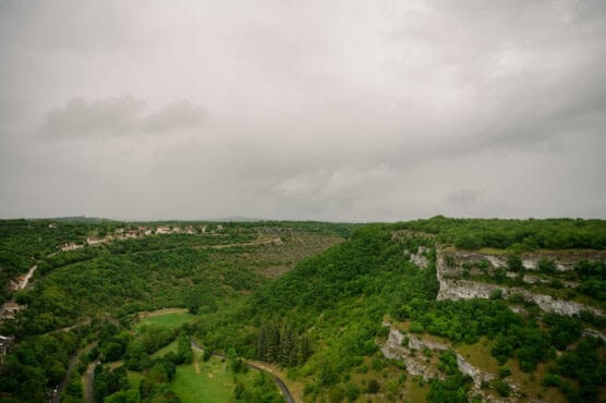 A lush, green valley stretches between rolling hills under a cloudy sky. Rocky cliffs with greenery are on the right, and houses are scattered along the ridge on the left. A road winds through the landscape below. by Justin Salem Meyer