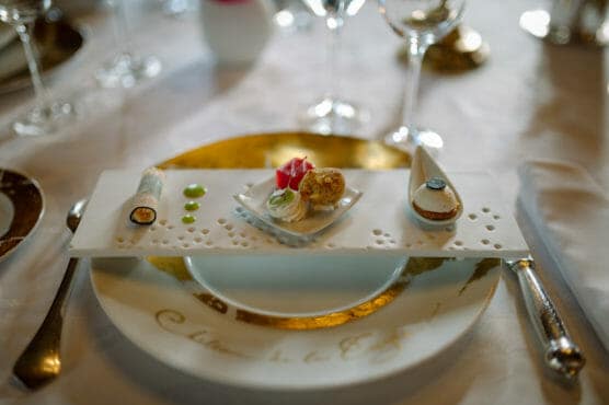 A gourmet appetizer trio is artfully arranged on a white plate with gold accents, featuring a rolled item, a small round bite, and a shell-shaped dish, surrounded by elegant tableware and wine glasses. by Justin Salem Meyer