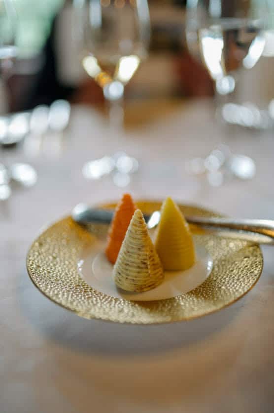 A decorative plate with three cone-shaped butters—one yellow, one orange, and one speckled—with a butter knife, set on a white tablecloth with wine glasses in the blurred background. by Justin Salem Meyer