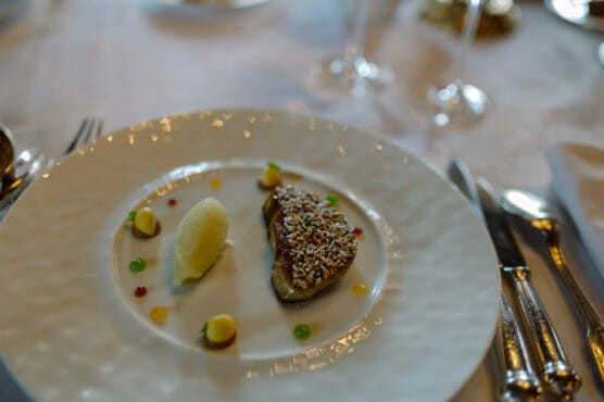 A gourmet dish on a white plate features a piece of meat topped with seeds, a scoop of pale yellow sorbet, and small colorful sauce dots, set on an elegant table with silver cutlery and wine glasses. by Justin Salem Meyer