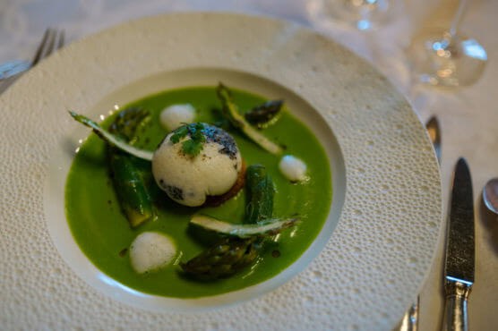 A gourmet dish featuring asparagus spears, a poached egg or mousse topped with foam, all served in a vibrant green sauce on a textured white plate. Tableware and glassware are visible around the plate. by Justin Salem Meyer