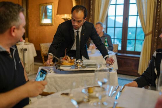 A waiter in a suit serves a dish on a tray to a table of diners in an elegant restaurant with large windows, white tablecloths, and warm lighting. One diner holds a phone, possibly taking a photo. by Justin Salem Meyer