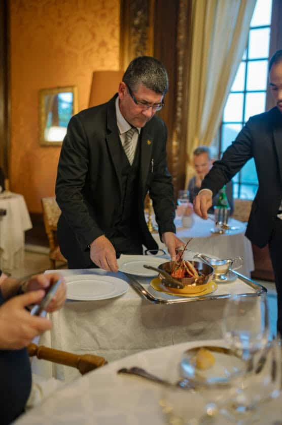 A waiter in a formal suit serves a lobster dish on a tray at an elegant restaurant, while another server assists. Diners sit at a table set with plates, glassware, and a bread roll. The room has ornate decor and large windows. by Justin Salem Meyer