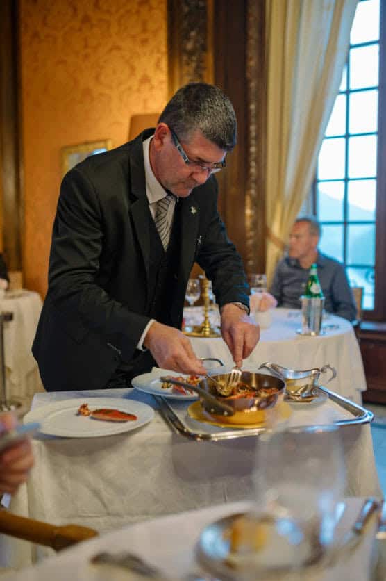 A waiter in a black suit prepares a dish at a table in an elegant restaurant with ornate decor. Other diners are seated at tables in the background near a large window. by Justin Salem Meyer