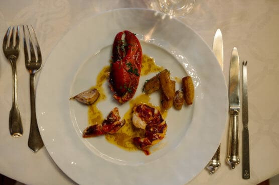 A white plate with lobster claw, lobster pieces in sauce, roasted potatoes, and garlic, set on a white tablecloth with a fork, knife, and spoon on either side of the plate. by Justin Salem Meyer