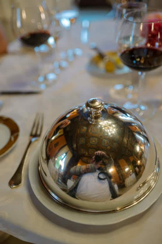A shiny silver cloche on a white plate reflects the photographer and a decorative ceiling. Wine glasses and plates are set on a white tablecloth, creating an elegant dining scene. by Justin Salem Meyer
