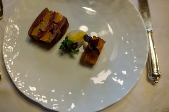 A white plate with a small, artfully arranged dish featuring slices of meat with sauce, a rectangular piece of fried food, a small dollop of yellow puree, and a garnish of herbs. A knife is placed to the right of the plate. by Justin Salem Meyer