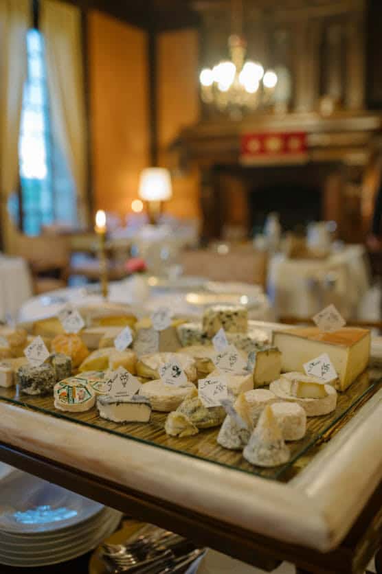 A table covered with a variety of cheeses, each labeled, in a warmly lit, elegant dining room with wooden paneling and chandeliers in the background. by Justin Salem Meyer