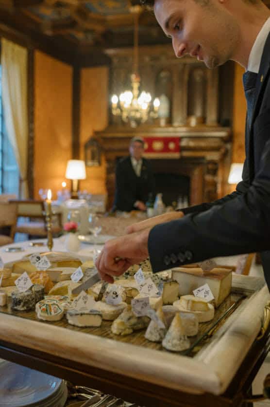 A waiter in a suit presents a tray filled with various cheeses labeled with small flags in an elegant, warmly lit dining room. A formally dressed person stands in the background near a fireplace. by Justin Salem Meyer