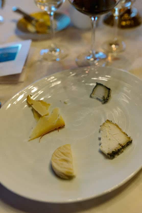 A white plate with four pieces of assorted cheese, each with different shapes and colors, sits on a table with two wine glasses and other dining items blurred in the background. by Justin Salem Meyer