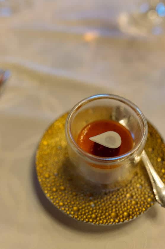 A small glass cup with a red sauce, topped with a white garnish, sits on a textured gold plate with a spoon beside it, on a white tablecloth. by Justin Salem Meyer