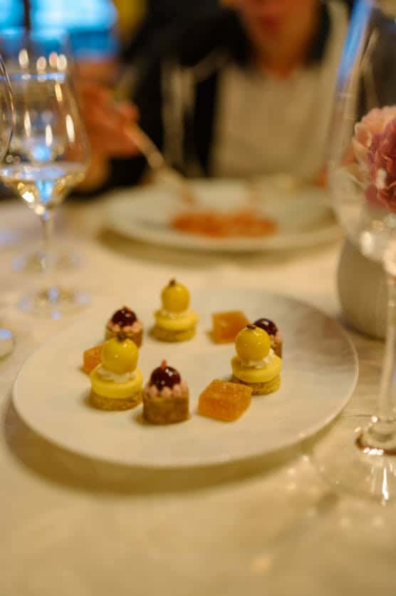 A white plate with assorted bite-sized gourmet desserts, including round yellow pastries and jelly cubes, set on an elegant table with wine glasses and blurred person in the background. by Justin Salem Meyer