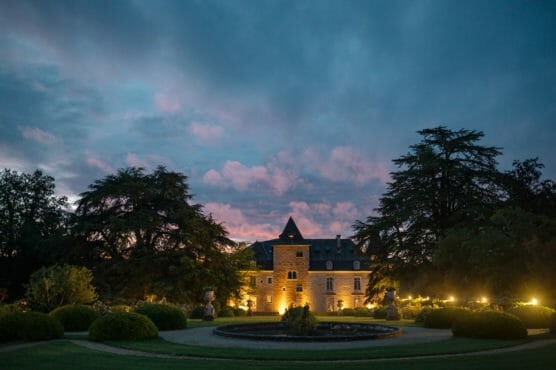 A grand stone chateau with a central tower is illuminated at dusk, surrounded by trees, manicured gardens, and a circular fountain under a dramatic, cloudy sunset sky. by Justin Salem Meyer