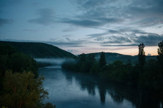 A serene river flows between tree-covered hills at dusk, with mist rising above the water and a dramatic, cloudy sky overhead as the sun sets in the distance. by Justin Salem Meyer