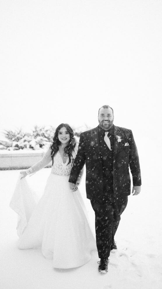 A bride in a long-sleeved white gown and a groom in a dark suit walk hand in hand through falling snow, smiling, with a snowy outdoor background. by Justin Salem Meyer