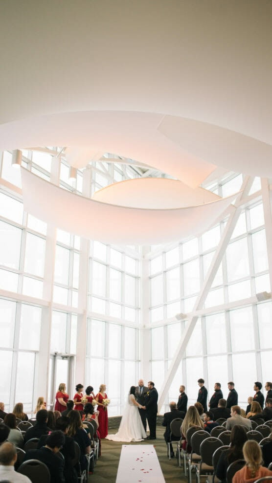 A bride and groom stand at the altar in a modern, airy venue with large windows, surrounded by their wedding party and seated guests watching the ceremony. White drapery hangs from the high ceiling above. by Justin Salem Meyer