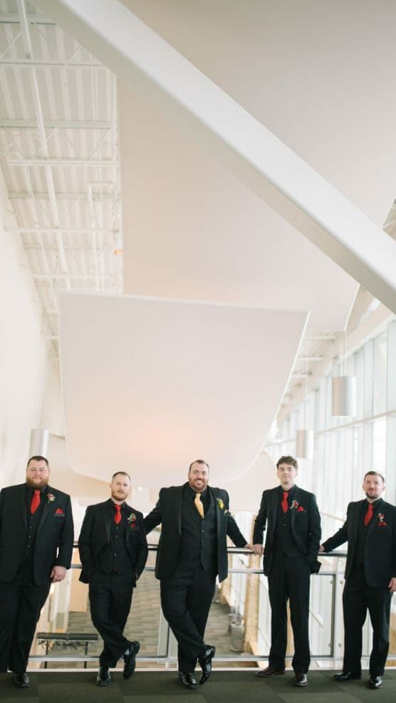Five men in black suits with red and yellow accents stand casually against a railing in a modern, bright building with high ceilings and large windows. They appear to be part of a wedding party. by Justin Salem Meyer