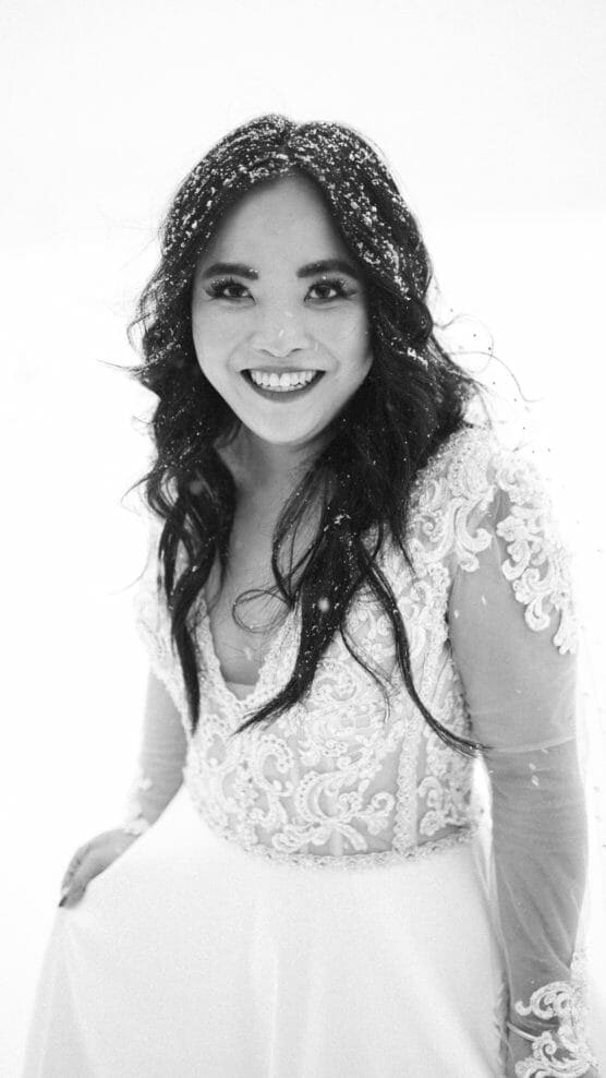 A woman in a lace wedding dress smiles at the camera, her dark hair covered with snowflakes. The photo is in black and white with a bright, snowy background. by Justin Salem Meyer