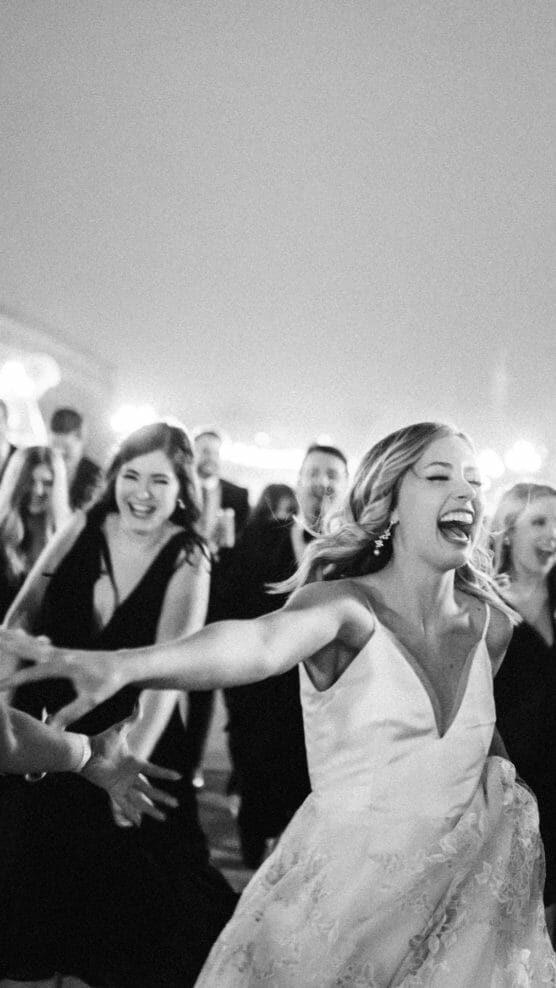 A joyful bride in a white dress laughs and reaches out while dancing, surrounded by friends in dark attire, during a lively celebration at night. The image is in black and white. by Justin Salem Meyer