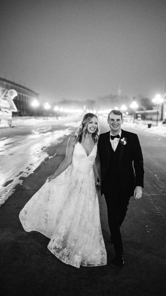 A bride in a white gown and a groom in a tuxedo walk hand in hand, smiling at night on a city street with lights and buildings in the background. The image is black and white. by Justin Salem Meyer