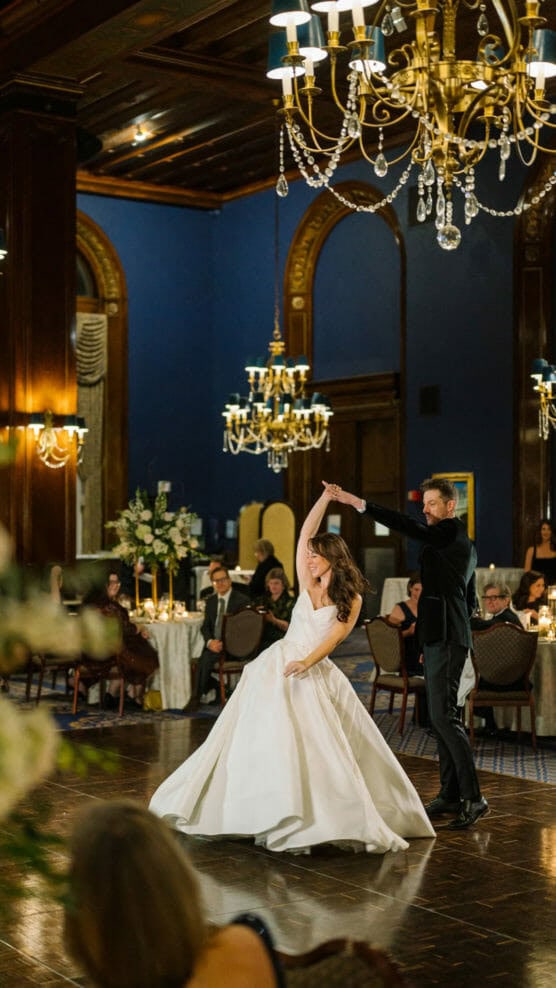 A bride in a white gown and a groom in a tuxedo dance elegantly under chandeliers in a grand, dimly lit ballroom, as guests seated at decorated tables watch them. by Justin Salem Meyer