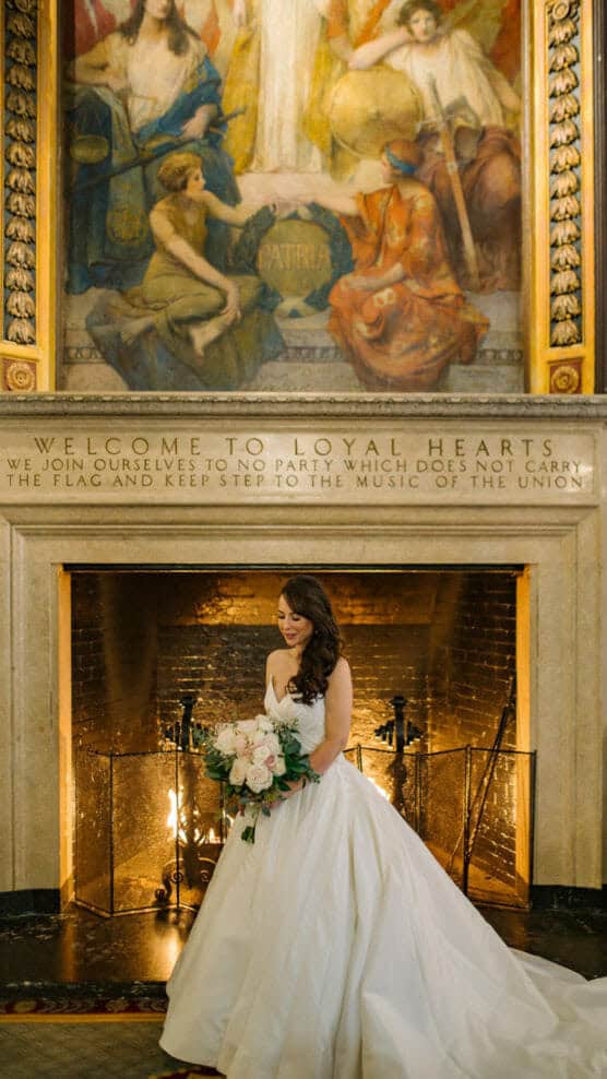 A bride in a white gown stands holding a bouquet in front of a large fireplace with an ornate mural and an inscription above it, in an elegant, historic room. by Justin Salem Meyer