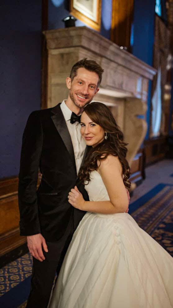 A man in a black tuxedo and a woman in a white wedding dress smile and pose together indoors in a warmly lit, elegant room with wood paneling and a large stone fireplace. by Justin Salem Meyer