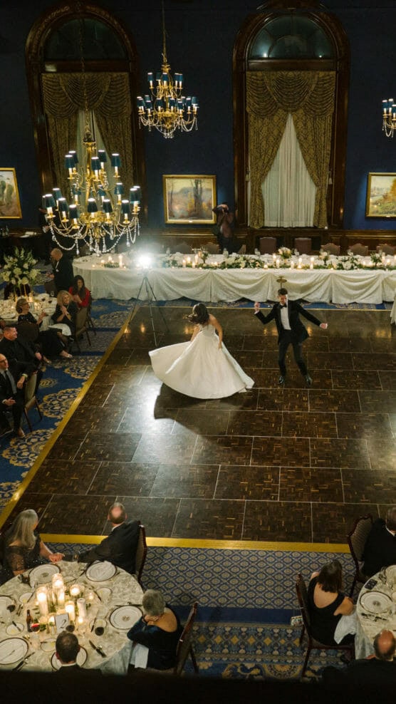 A bride in a white gown and a groom in a tuxedo dance on a large wooden floor in an elegant, chandelier-lit ballroom, surrounded by seated guests at round tables. by Justin Salem Meyer