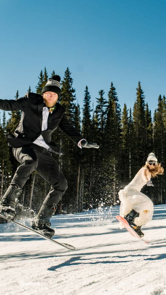 A man in a suit and a woman in a wedding dress snowboard down a snowy slope surrounded by pine trees, both wearing winter hats and gloves, with snow spraying as they ride. by Justin Salem Meyer
