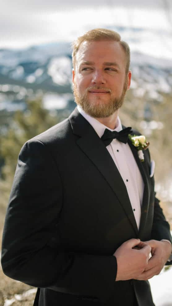 A groom in a black tuxedo with a bow tie and boutonniere stands outdoors, looking off to the side with a slight smile. Snowy mountains and trees are visible in the background. by Justin Salem Meyer