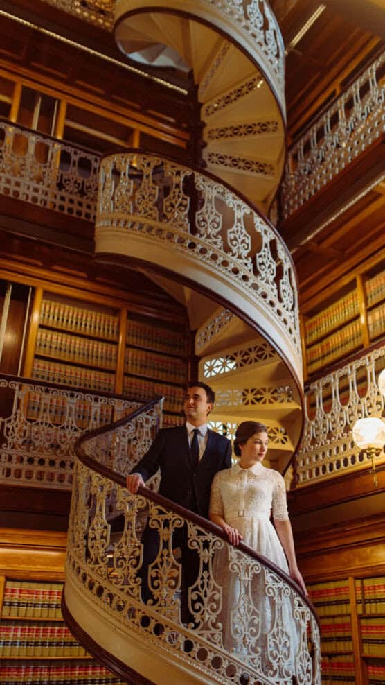 A couple stands on an ornate spiral staircase in a grand library lined with bookshelves. The man wears a suit and the woman wears a white lace dress. Warm light highlights the elegant ironwork and wood paneling. by Justin Salem Meyer