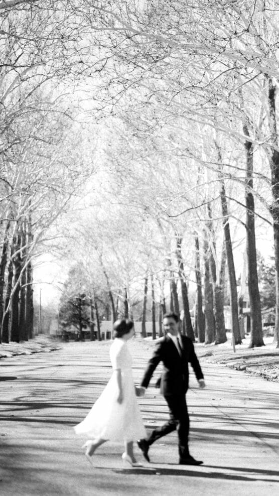 A black and white photo of a couple walking across a tree-lined street. The woman wears a light dress and the man wears a suit, both slightly blurred in motion, with tall leafless trees in the background. by Justin Salem Meyer