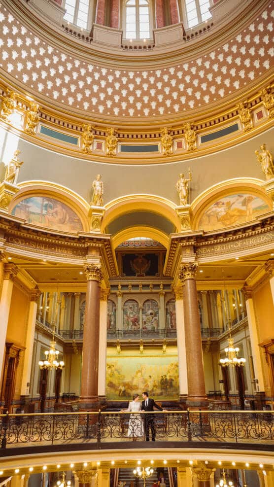 A couple stands on a balcony inside an ornate domed building with columns, arches, gold accents, and paintings, looking up at the detailed ceiling under soft, warm lighting. by Justin Salem Meyer