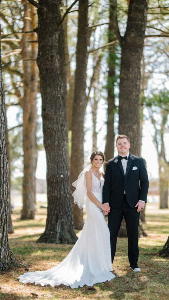 A bride in a white gown and veil stands holding hands with a groom in a black tuxedo, posing together among tall trees in a sunlit, wooded outdoor setting. by Justin Salem Meyer