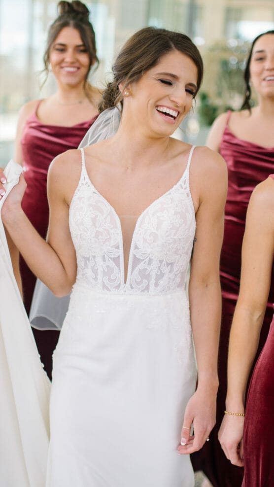 A smiling bride in a white lace wedding dress holds her gown, surrounded by bridesmaids in matching burgundy dresses. The group appears joyful, standing indoors with natural light. by Justin Salem Meyer